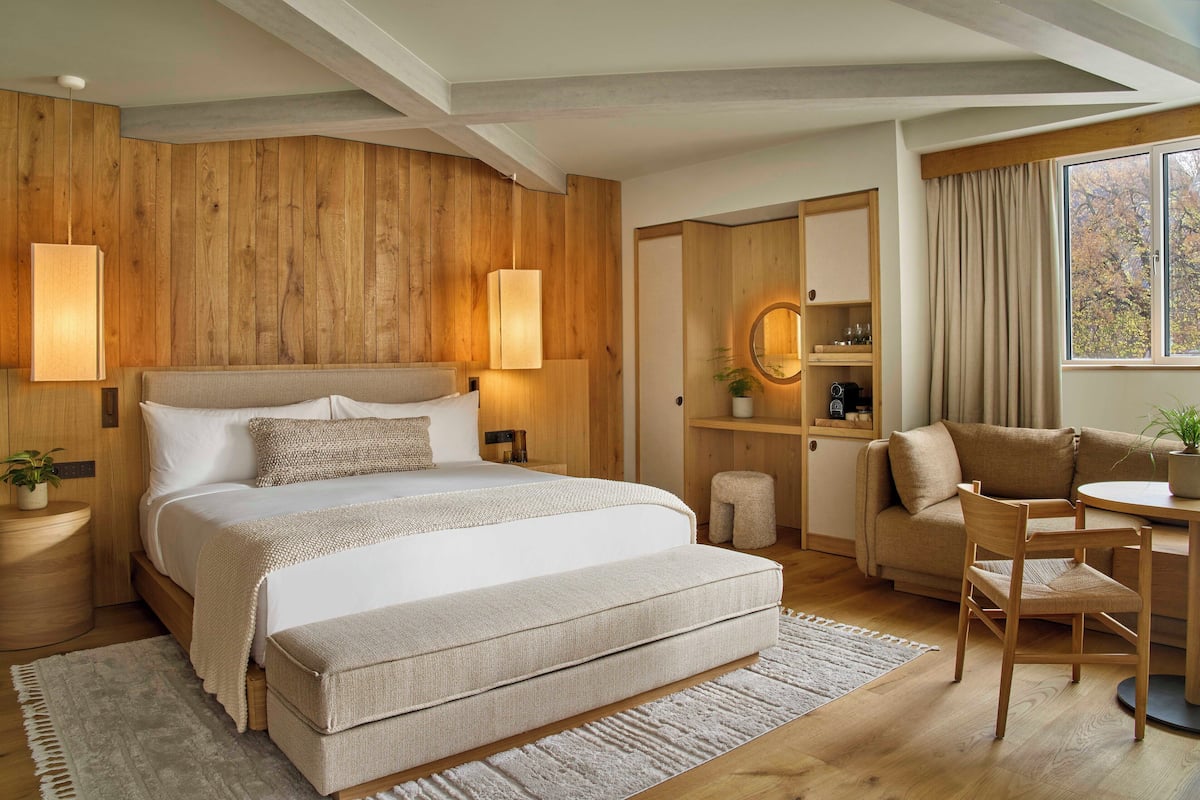 A modern bedroom with a large bed, bedside lamps, a wooden headboard, a small table with a chair, and a window with curtains.