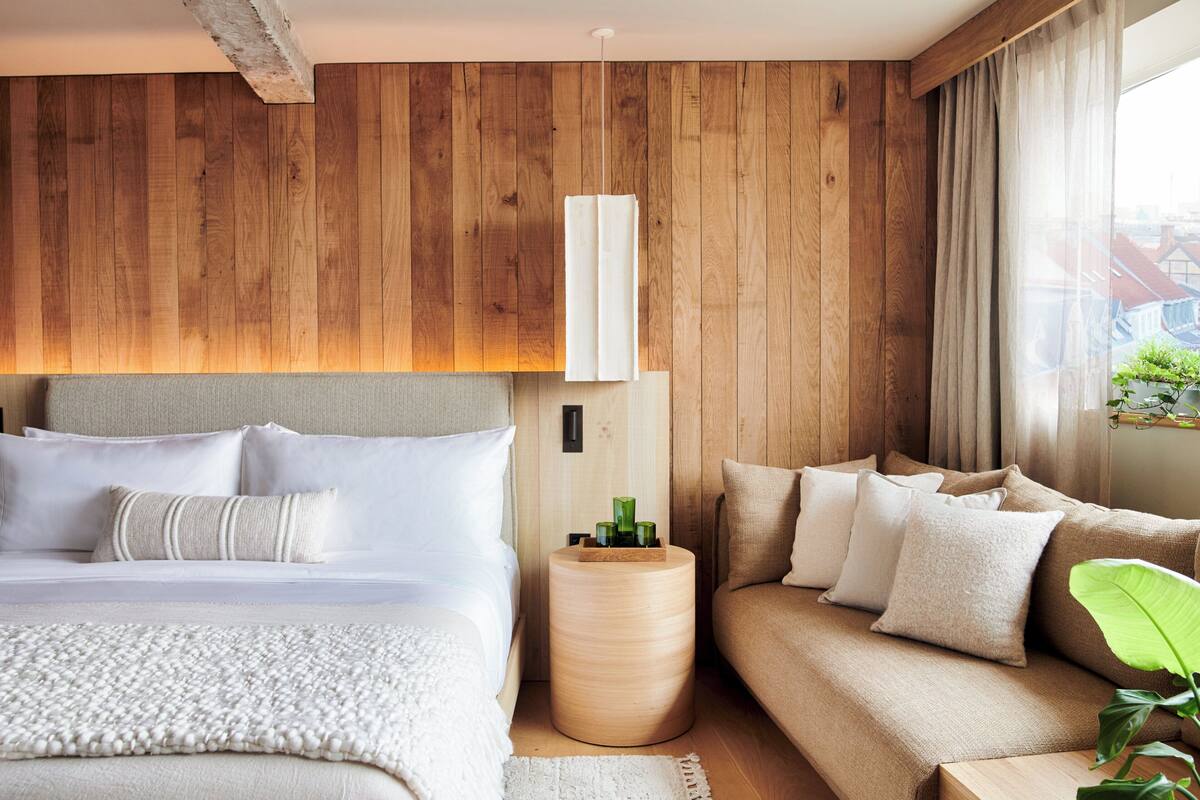 A bedroom with a wooden headboard, a bed with white bedding, a round wooden nightstand, and a beige sofa.
