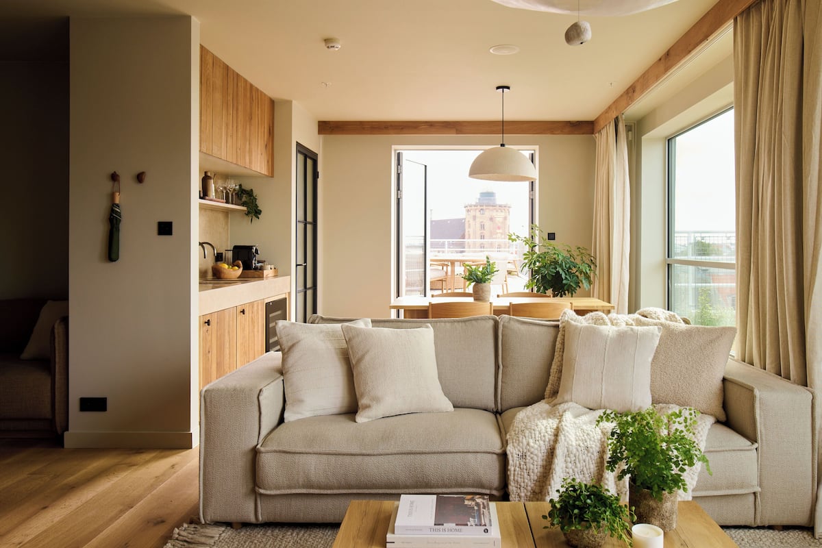 A modern living room with a beige sofa, wooden beams, a dining area, and large windows.