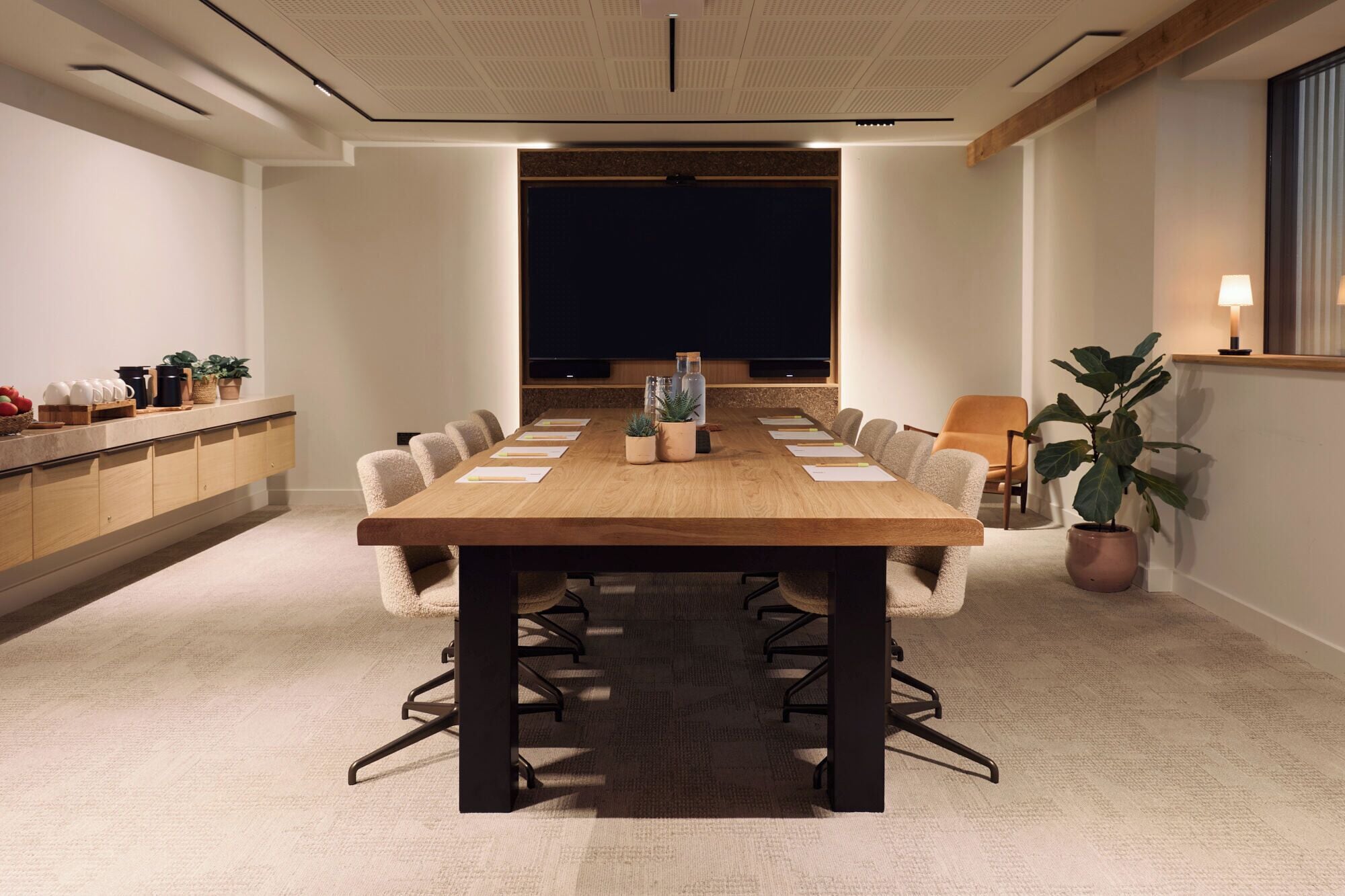 Un salón de conferencias con una larga mesa, sillas, una pantalla de proyector y una planta en maceta.