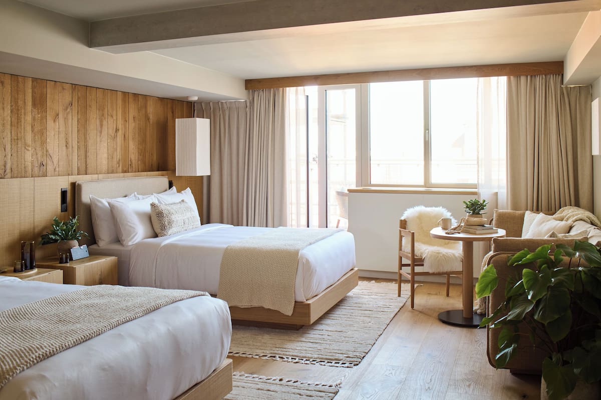 A hotel room with two beds, a small table, a chair, and a large window with curtains.