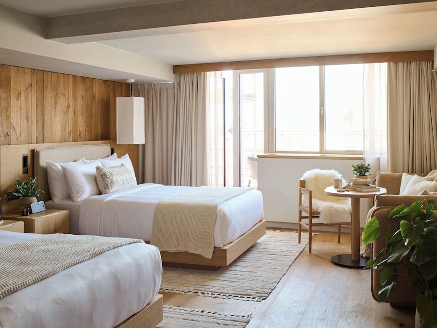 Een hotelkamer met twee bedden, een tafeltje, een stoel en een groot raam met gordijnen.