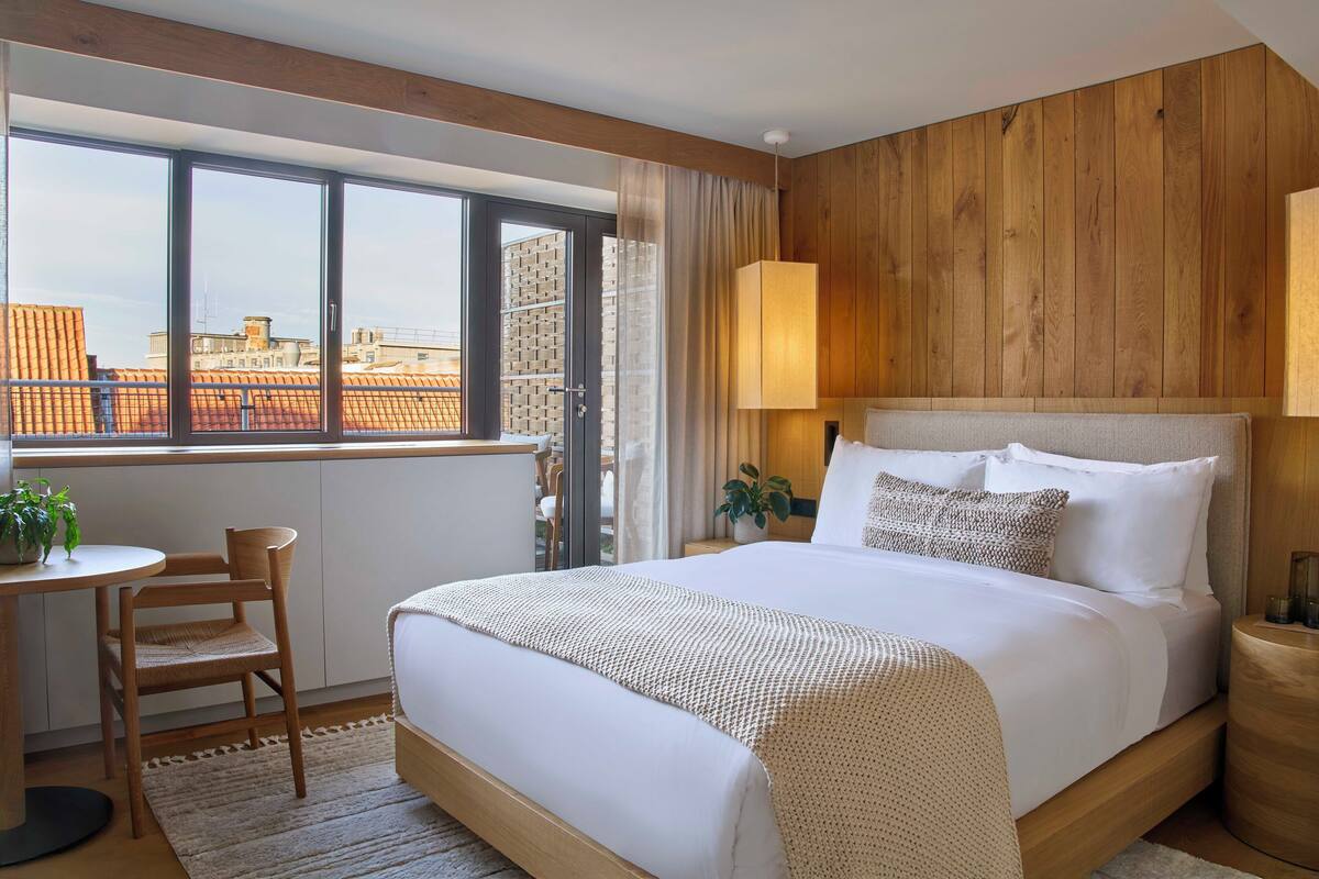 A hotel room with a large bed, a wooden headboard, a window with a view of buildings, a small table with a chair, and a bedside lamp.
