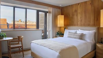 A modern hotel room with a large bed, a wooden headboard, a window with a city view, a small table with a chair, and a bedside lamp.
