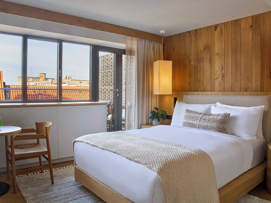 A modern hotel room with a large bed, a wooden headboard, a window with a city view, a small table with a chair, and a bedside lamp.