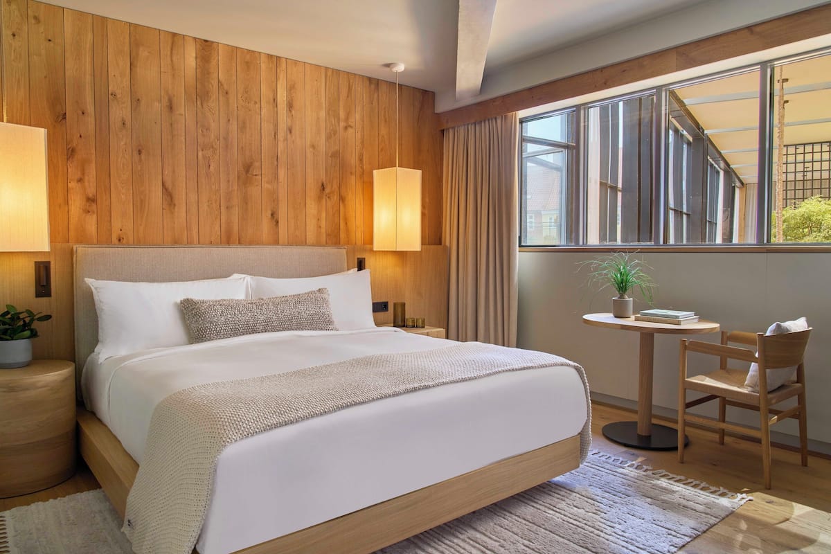 A hotel room with a wooden headboard, a large bed, a bedside table with a lamp, a small round table with a chair, and a window with curtains.