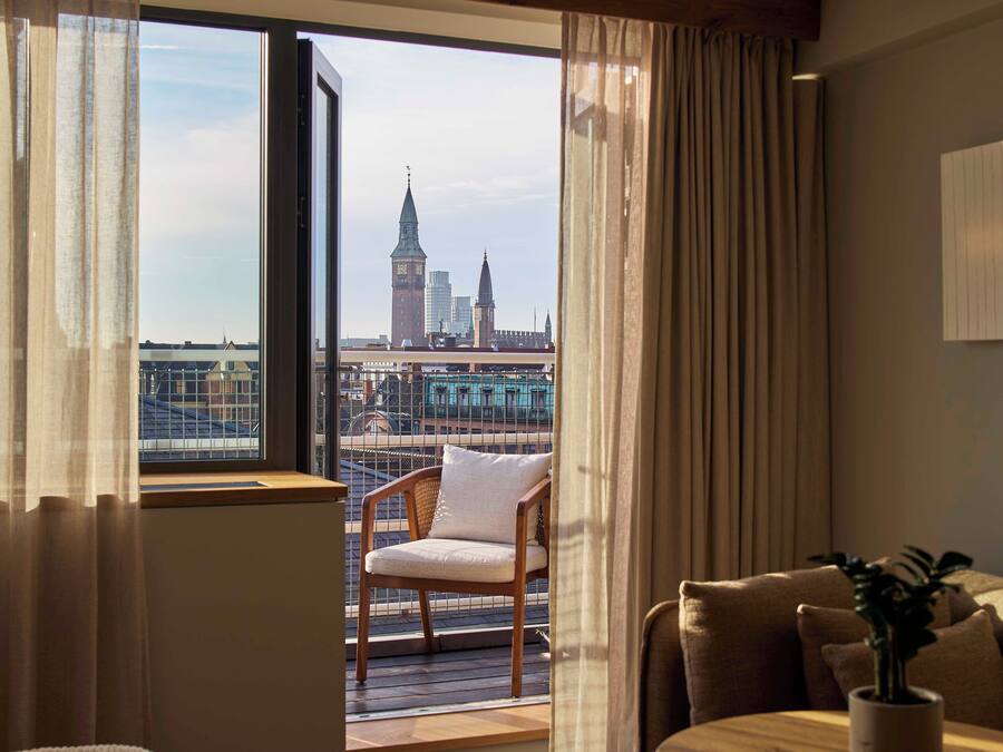 Et hotelværelse med balkon, en træstol, et spisebord og udsigt over bybilledet med bygninger og en bro.