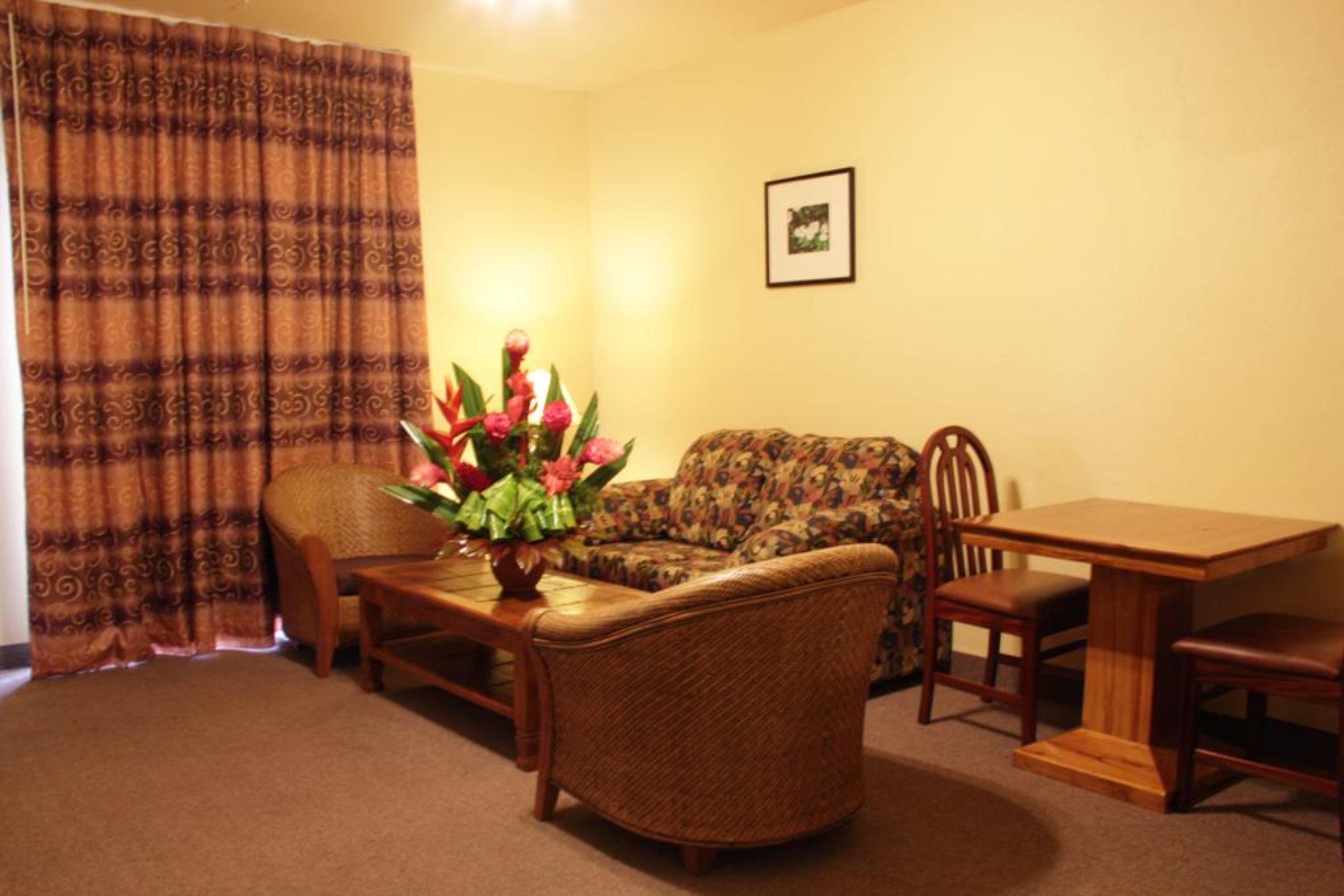 A living room with a sofa, a coffee table with a floral centerpiece, a dining table with chairs, and a curtain.