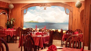 A restaurant with wooden walls, red tablecloths, and a mural of a coastal landscape. - Tradewinds Hotel (Ottoville)