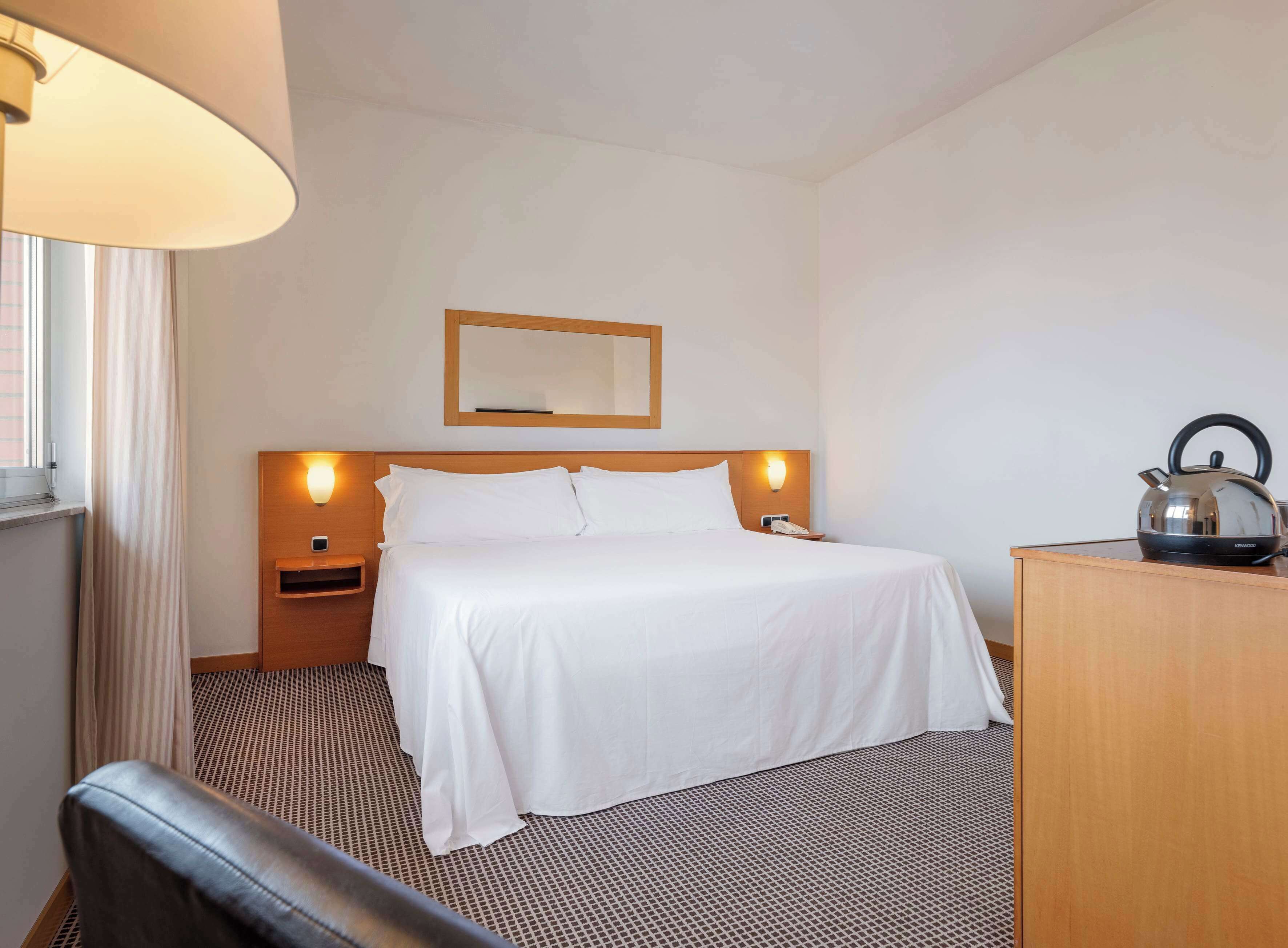 Quarto de hotel com uma cama grande, cabeceira de madeira, mesa de cabeceira com abajur, espelho sobre a cama, chaleira em um armário de madeira e uma cadeira.