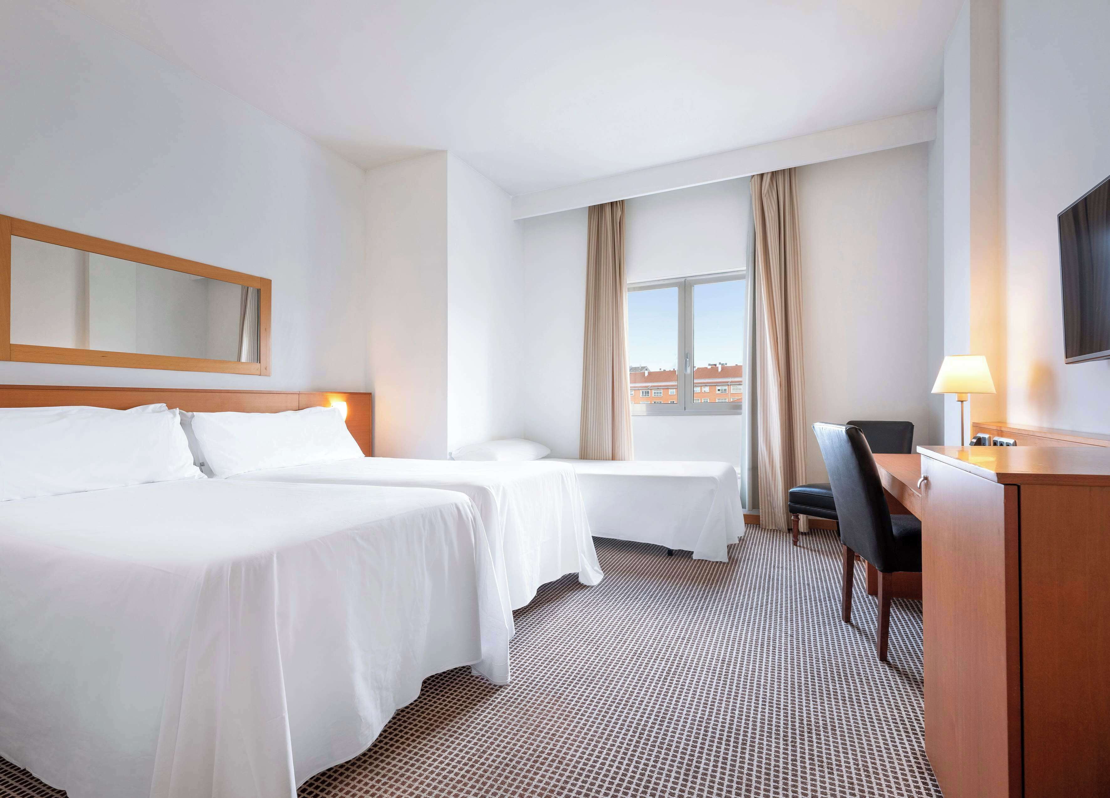 A hotel room with two beds, a desk, a chair, a TV, and a window with curtains.