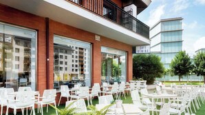 An outdoor cafe with white chairs and tables, surrounded by greenery and a modern building in the background.
