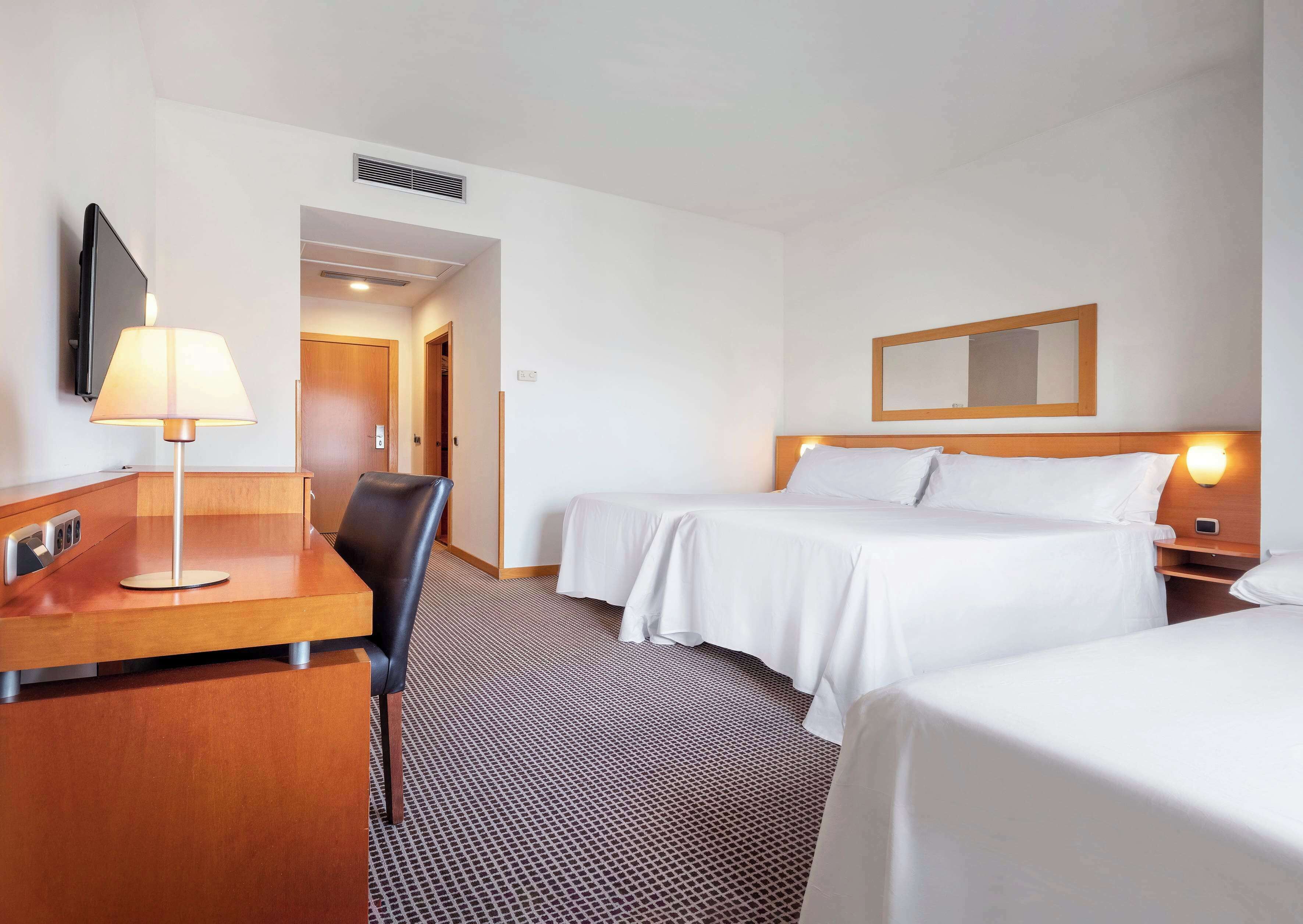 Habitación de hotel con escritorio, dos camas, una silla, una lámpara, un espejo y una luz empotrada en la pared.