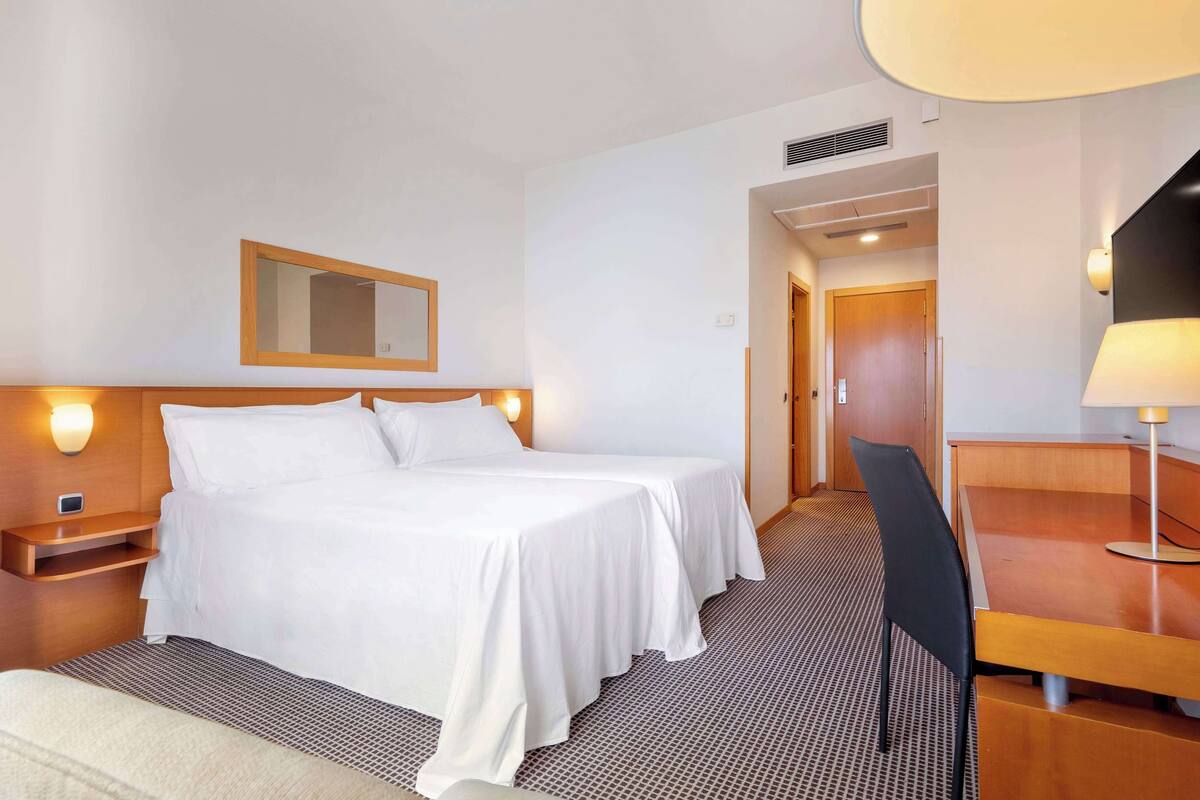 A hotel room with a double bed, bedside tables, a desk with a chair, a lamp, and a mirror on the wall.