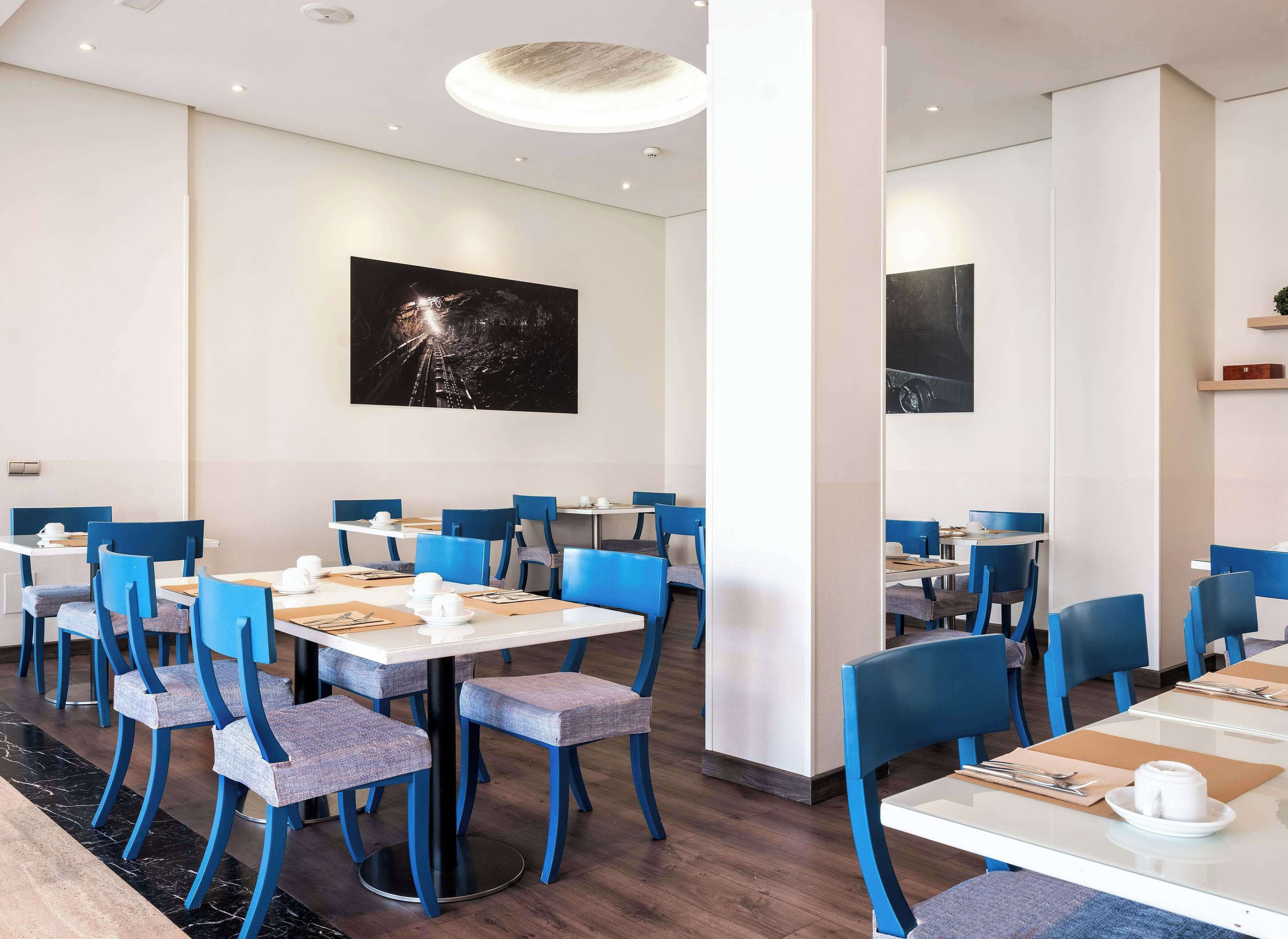 A modern dining area with blue chairs and white tables, featuring a large black and white artwork on the wall.