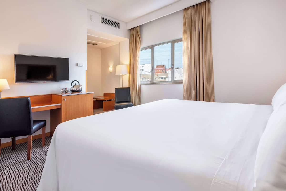 A hotel room with a large bed, a desk with a chair, a television, and a window with curtains.