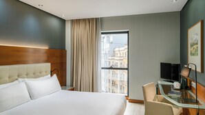 Et hotelværelse med en stor seng, et skrivebord med stol, et fjernsyn og et vindue med udsigt til bygninger.
