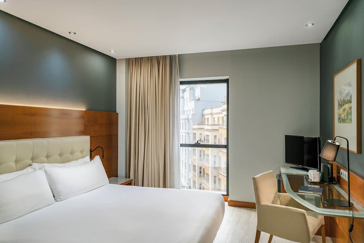 A hotel room with a large bed, a desk with a chair, a television, and a window with a view of buildings.