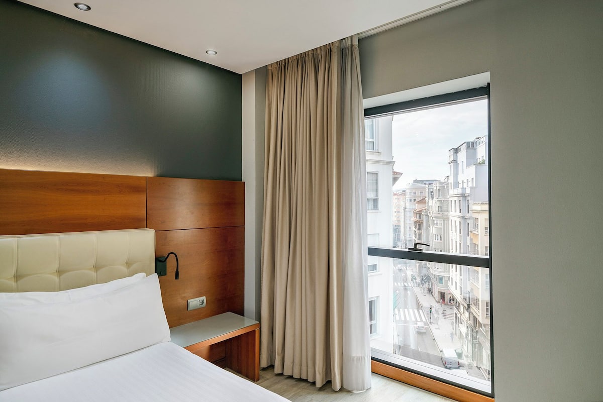 A hotel room with a large window, a wooden headboard, a bed, and a nightstand.