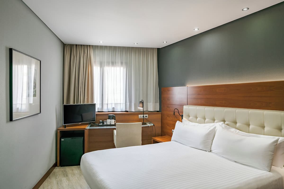 A hotel room with a bed, a desk with a computer, a chair, a television, and a window with curtains.