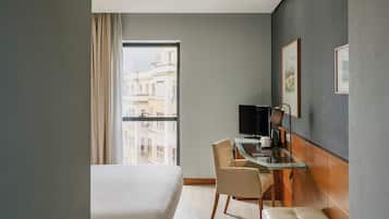 Quarto de hotel com uma cama grande, uma escrivaninha com computador, uma cadeira, um banco e vista da cidade pela janela.