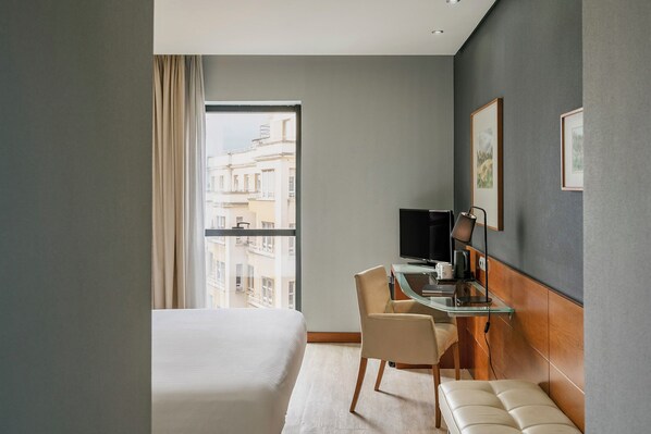 A hotel room with a large bed, a desk with a computer, a chair, a bench, and a view of the city through the window.