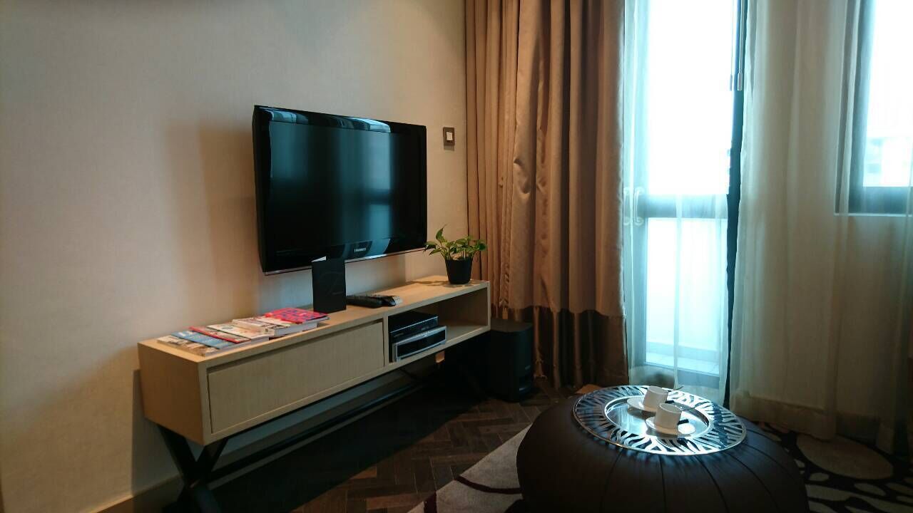 A modern hotel room with a flat-screen TV mounted on the wall, a small table with a potted plant, and a patterned ottoman.