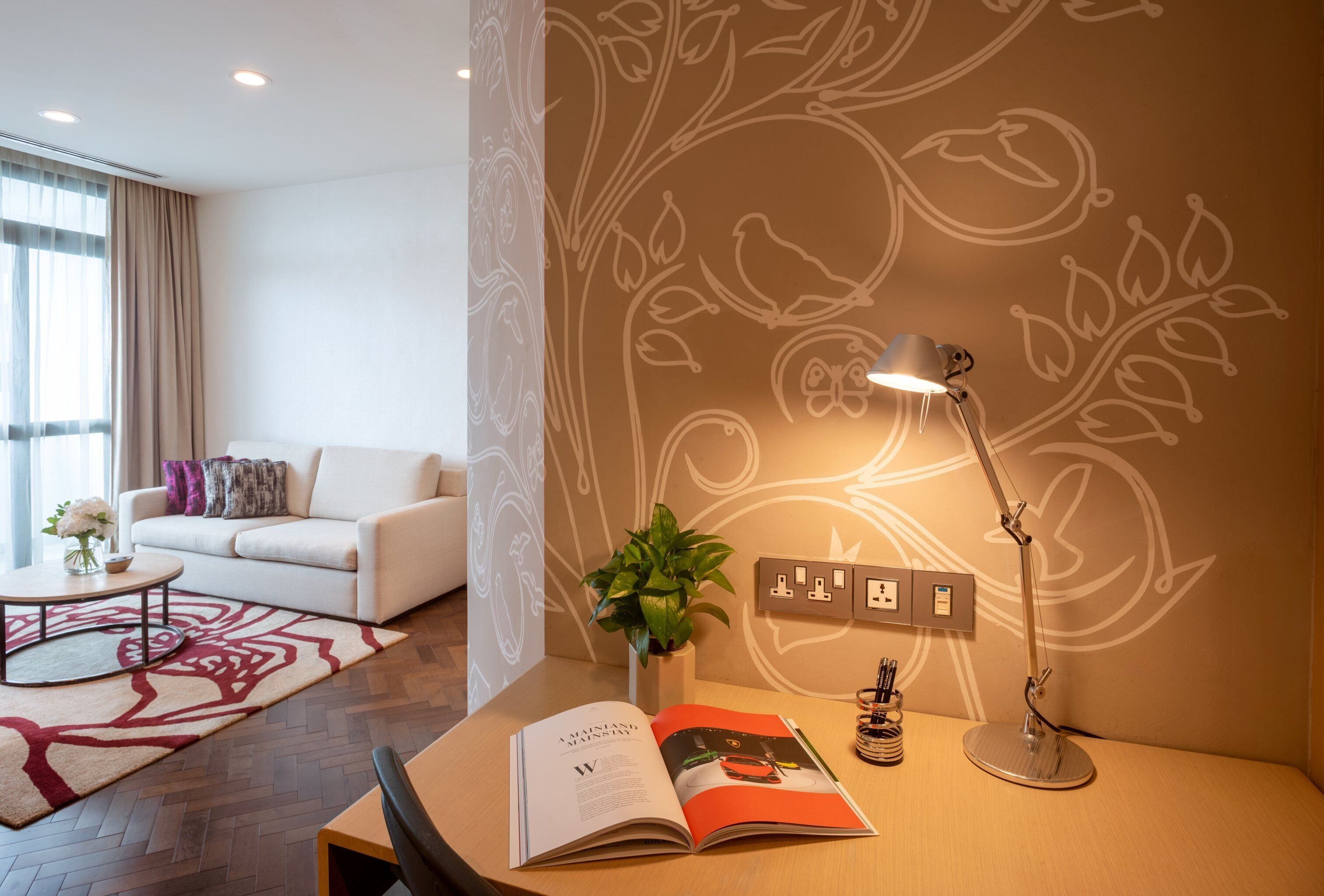 Un salon moderne avec un canapé blanc, une table basse surmontée d’un tapis à motifs floraux, un bureau avec une lampe, une plante et un magazine.