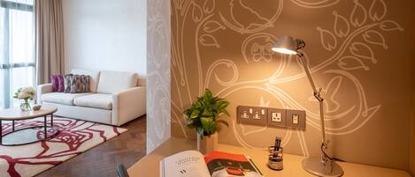 Un salon moderne avec un canapé blanc, une table basse surmontée d’un tapis à motifs floraux, un bureau avec une lampe, une plante et un magazine.