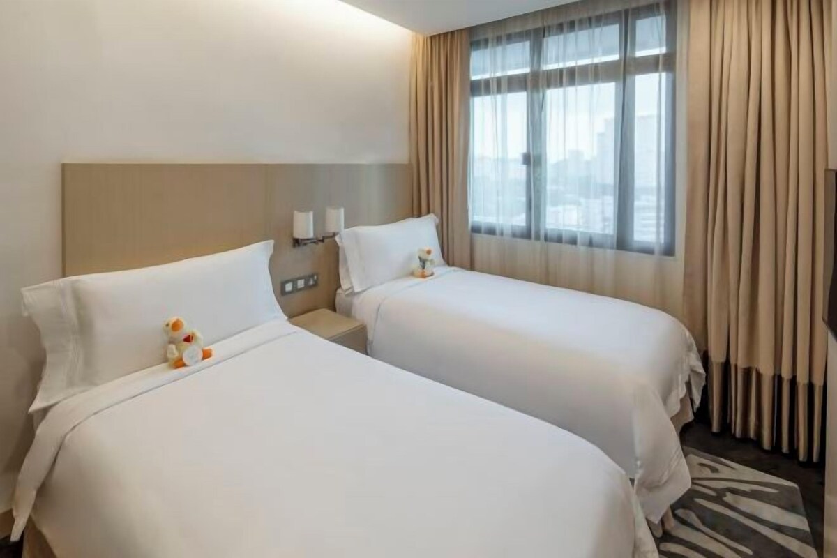 Habitación de hotel con dos camas, cabecero, lámparas de noche, ventana con cortinas y vista a la ciudad.
