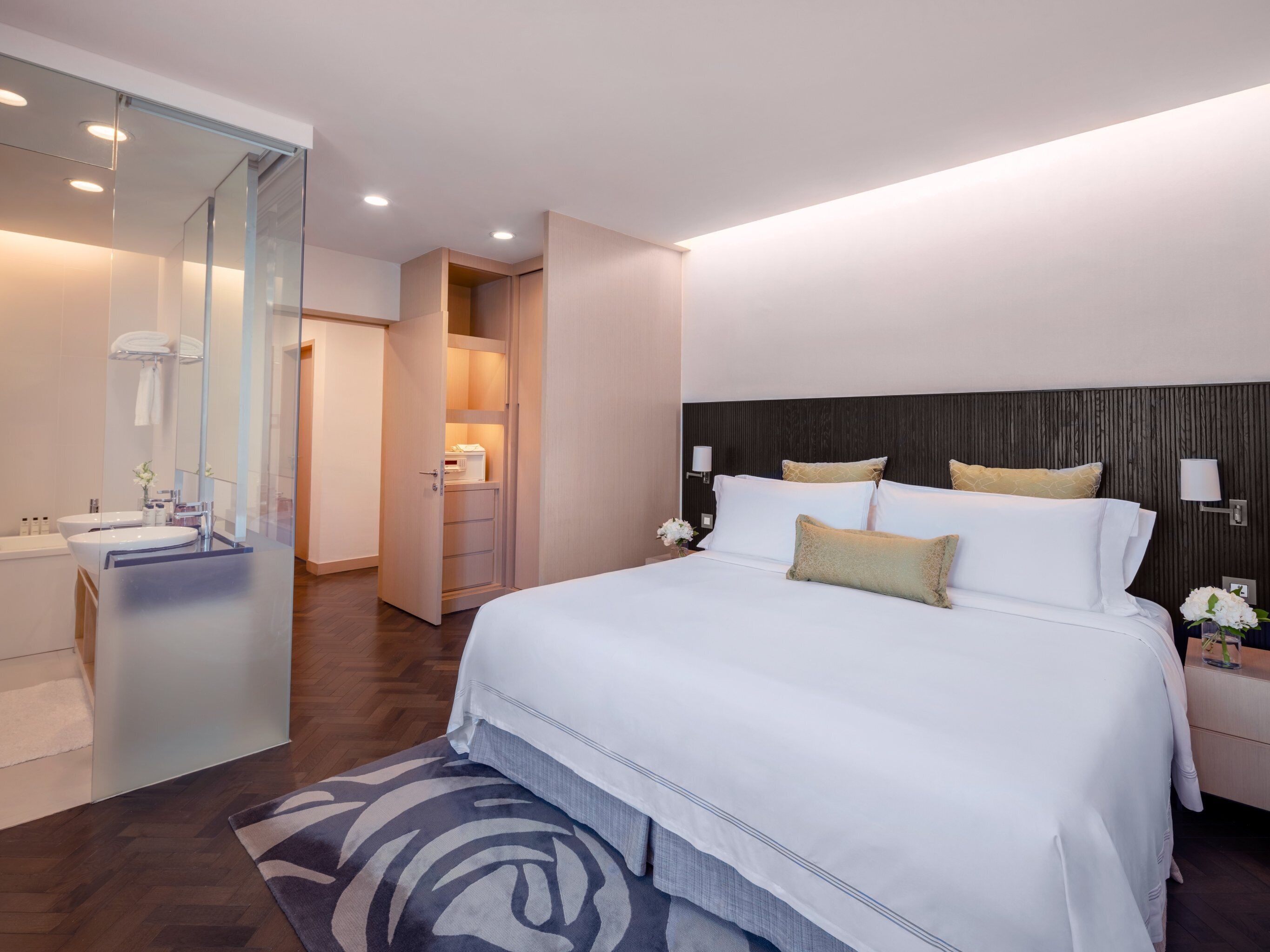 A modern hotel room with a large bed, a walk-in closet, a bathroom with a glass partition, and a wooden floor.