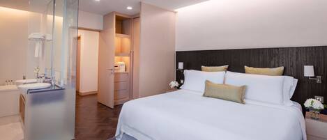 A modern hotel room with a large bed, a walk-in closet, a bathroom with a glass partition, and a wooden floor.