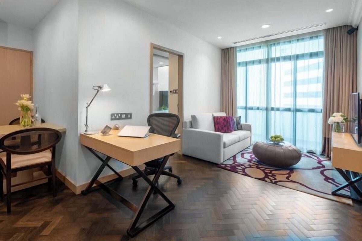 Una habitación de hotel moderna con piso de madera en patrón espiga, un escritorio con silla, un sofá, un ventanal con cortinas y un tapete decorativo.