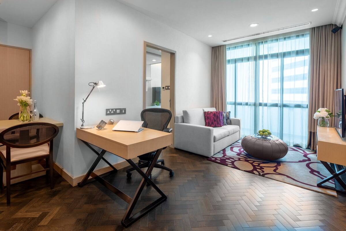 A modern hotel room with a wooden herringbone floor, a desk with a lamp, a sofa, a coffee table, and a large window with curtains.