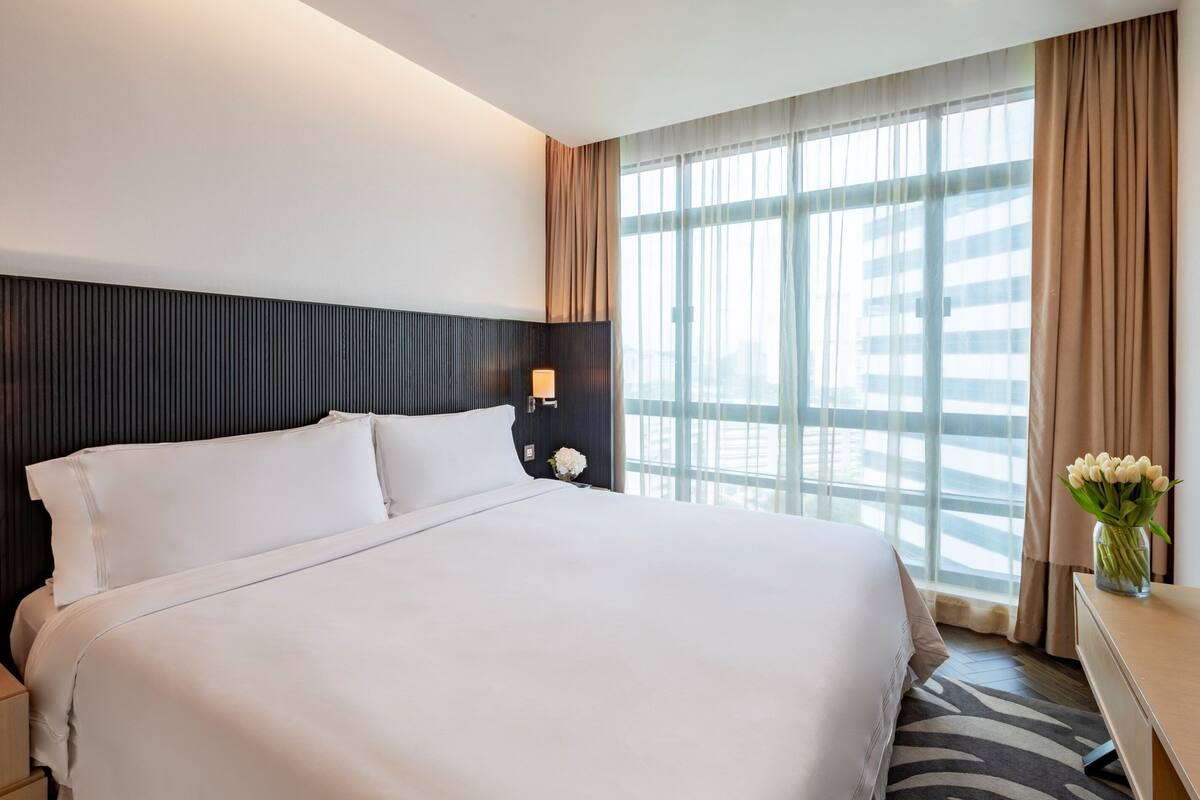 A hotel room with a large bed, a nightstand with a lamp, a vase with flowers, and a view of the city through large windows.