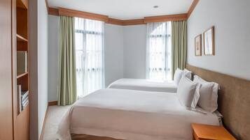 A hotel room with two beds, a wooden nightstand, a bookshelf, and large windows with sheer curtains.