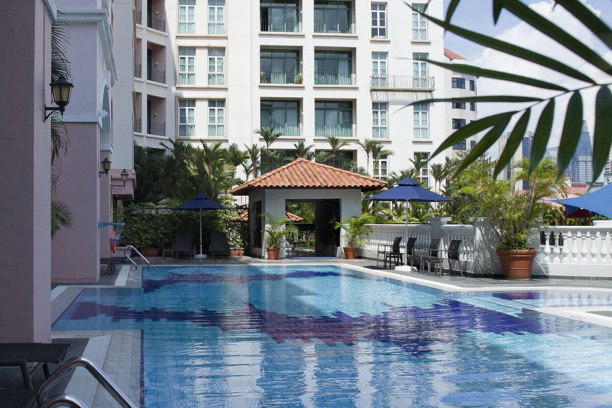 A swimming pool with a clear blue water surface, surrounded by a building with multiple balconies and a tiled roof.