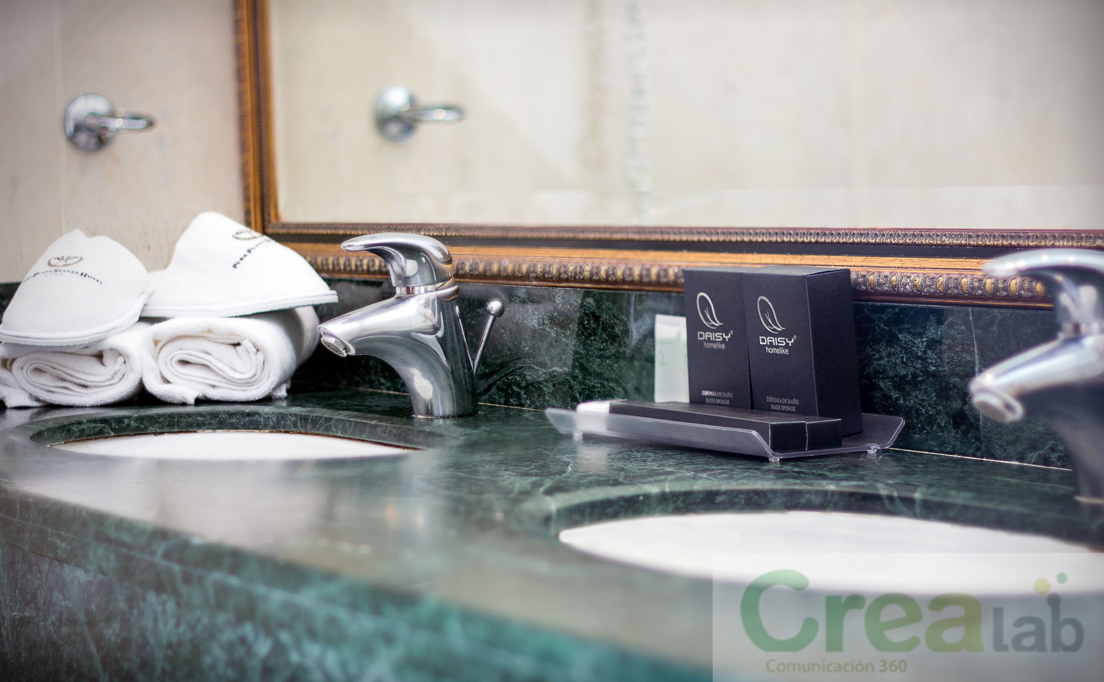 A bathroom with two sinks, a mirror, and a towel rack with folded towels. There are two boxes on the counter, one labeled "DAISY" and the other "CREALAB".