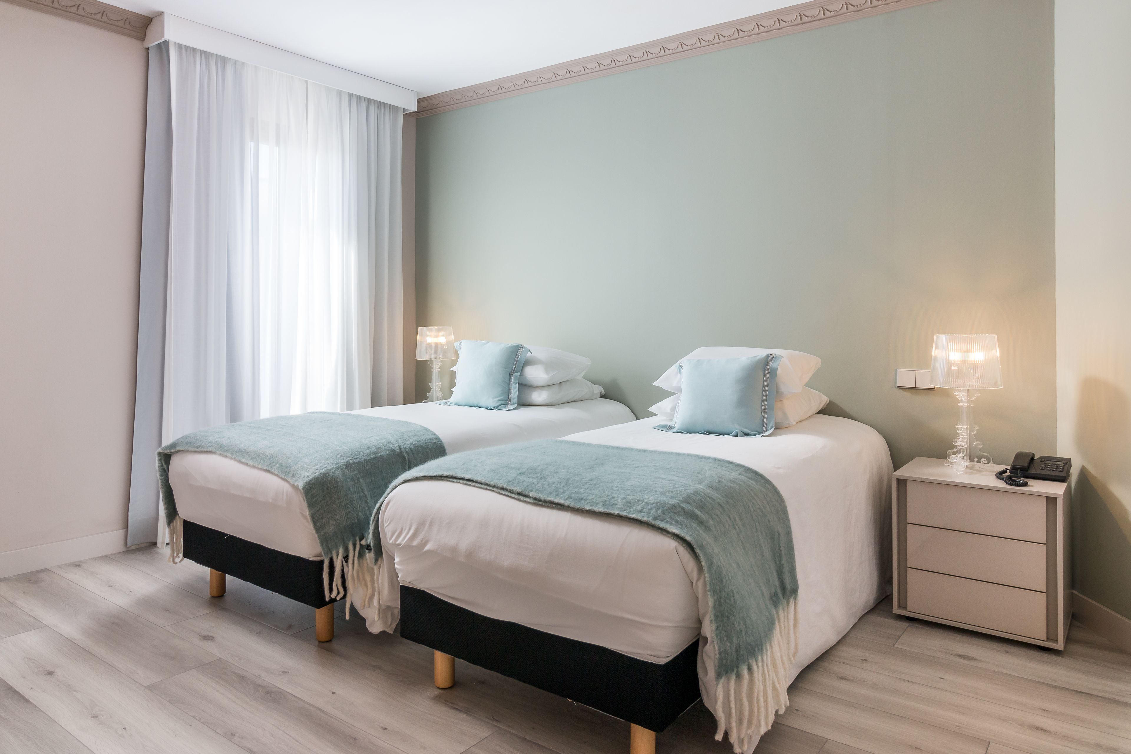Une chambre d’hôtel avec deux lits, chacun doté d’une tête de lit noire et blanche, une table de chevet avec une lampe et une fenêtre ornée de rideaux légers.