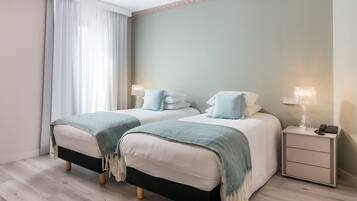 Une chambre d’hôtel avec deux lits, chacun doté d’une tête de lit noire et blanche, une table de chevet avec une lampe et une fenêtre ornée de rideaux légers.
