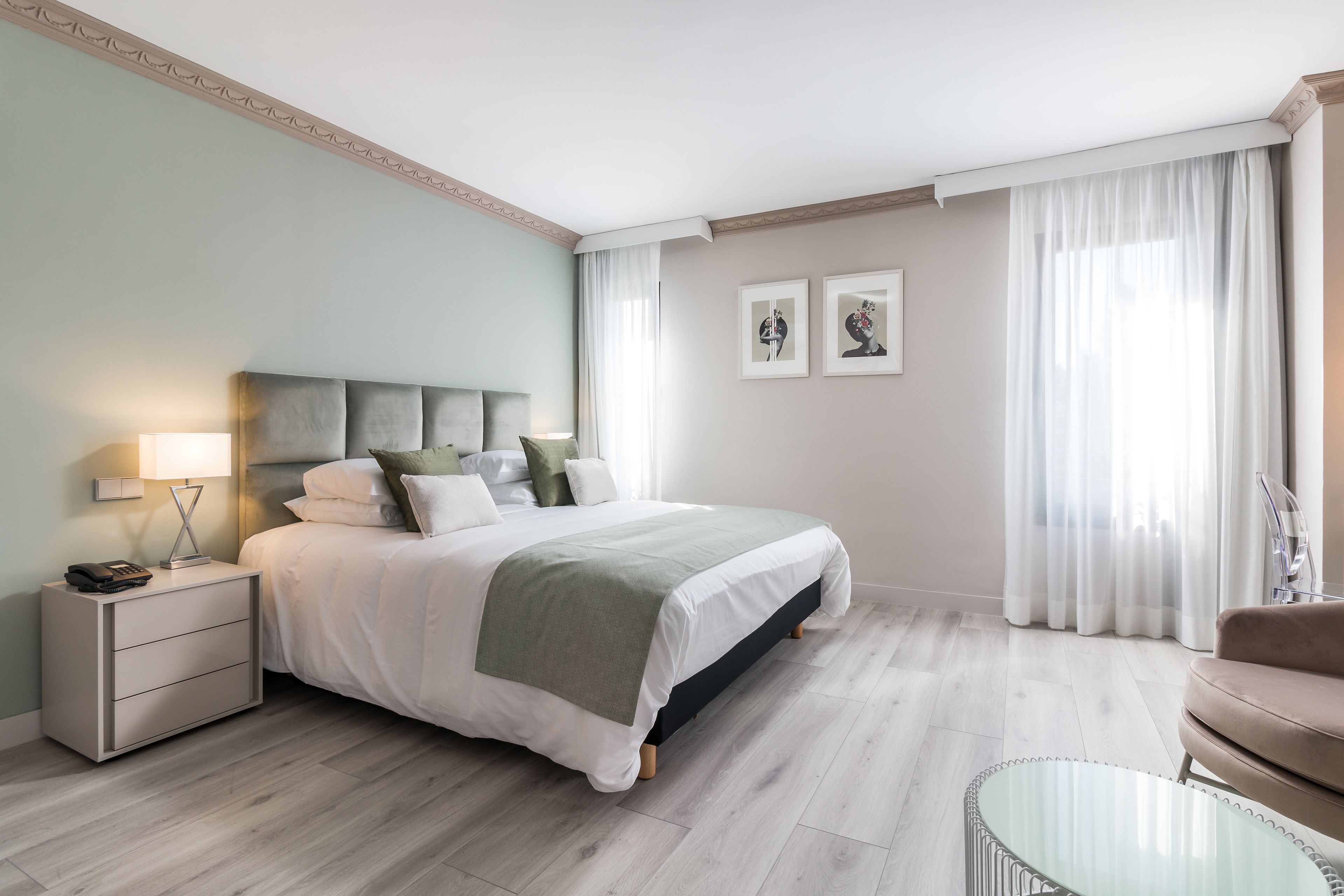 Habitación de hotel moderna con una cama bien hecha, una mesita de noche con una lámpara, una mesa de centro de cristal y vistas a la ciudad a través de amplias ventanas.