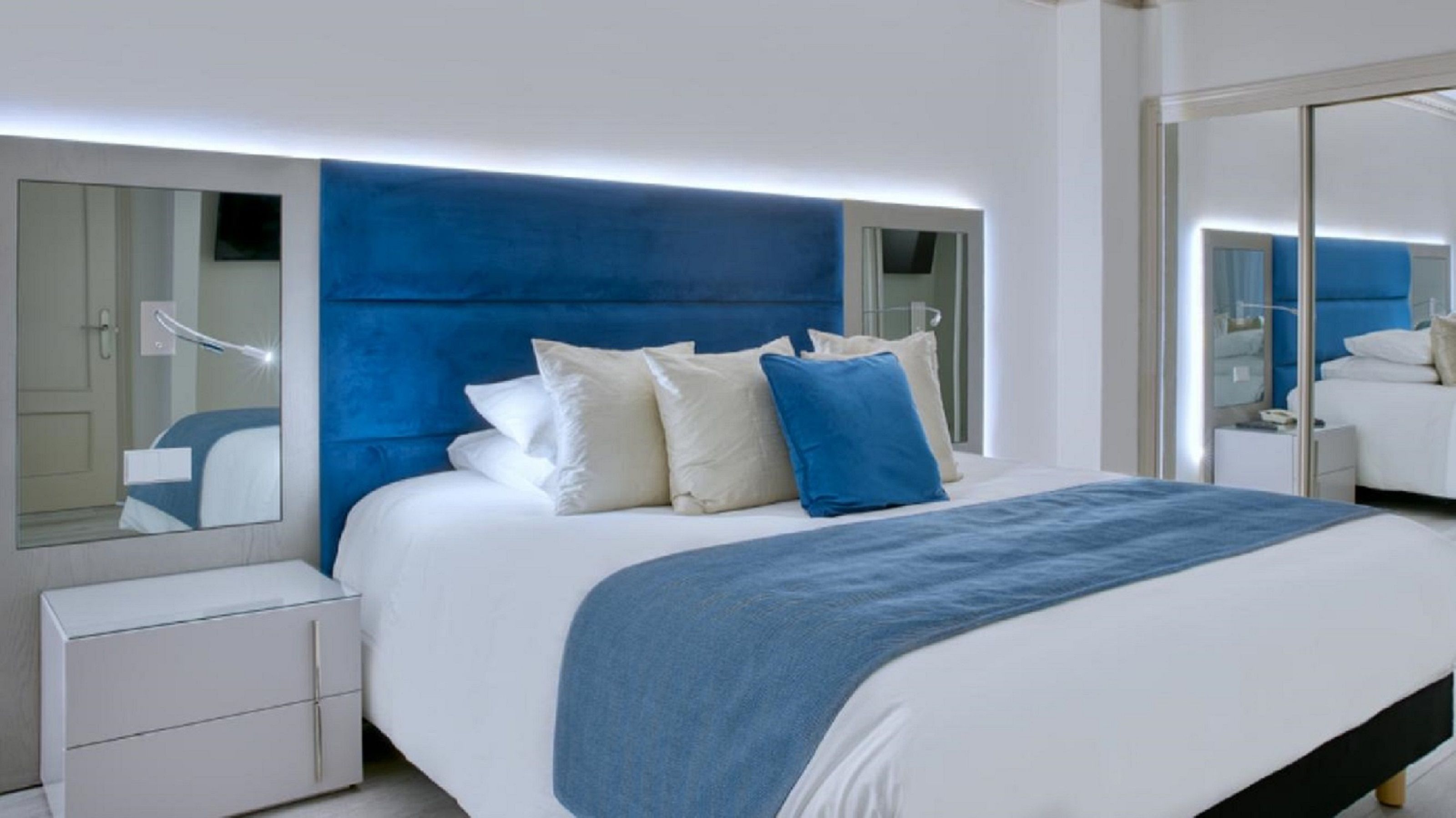 A modern hotel room with a large bed, a bedside table, a mirror, and a wardrobe.