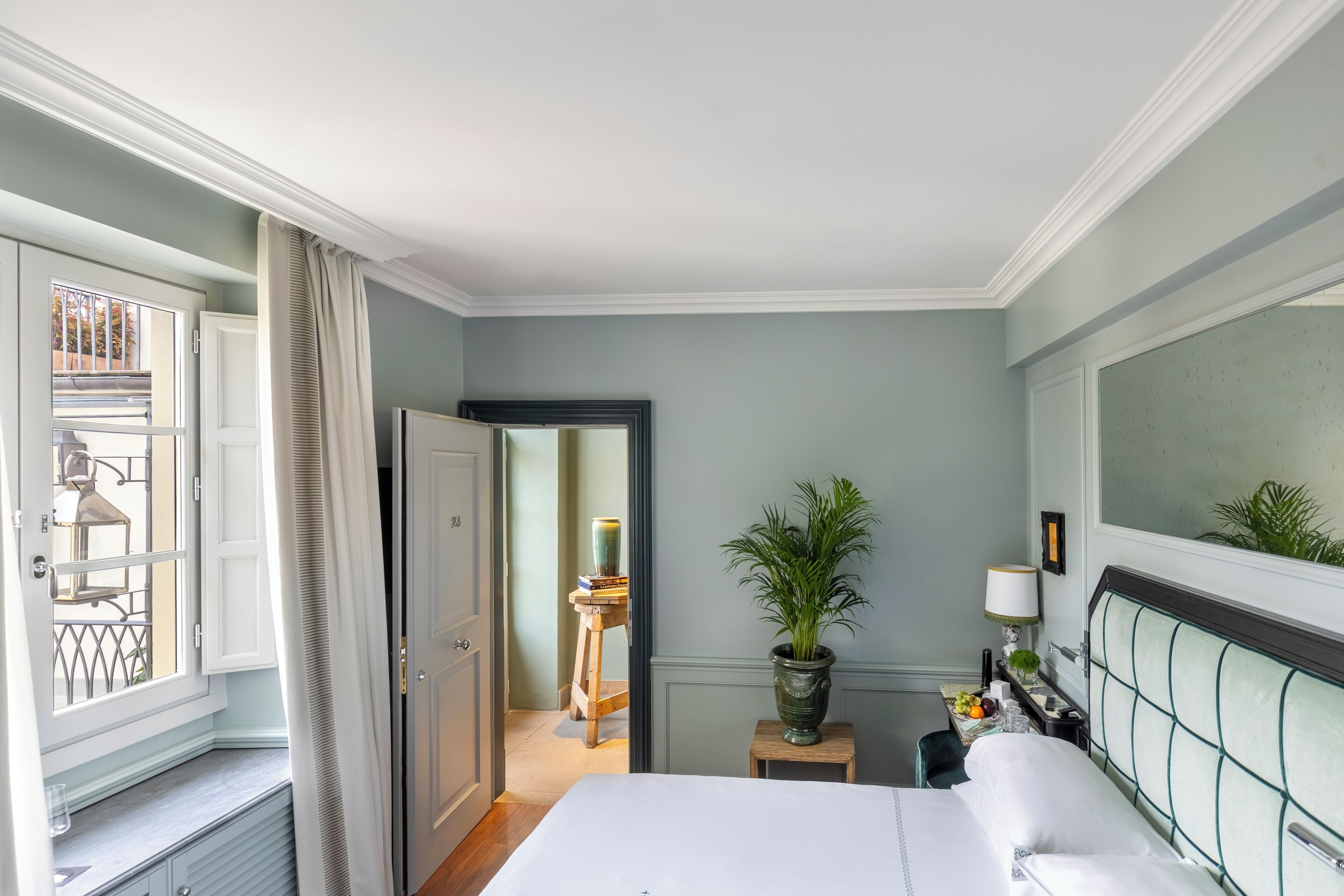 Habitación de hotel con cama, mesita de noche, planta en maceta, ventana con cortinas y vista al balcón exterior.