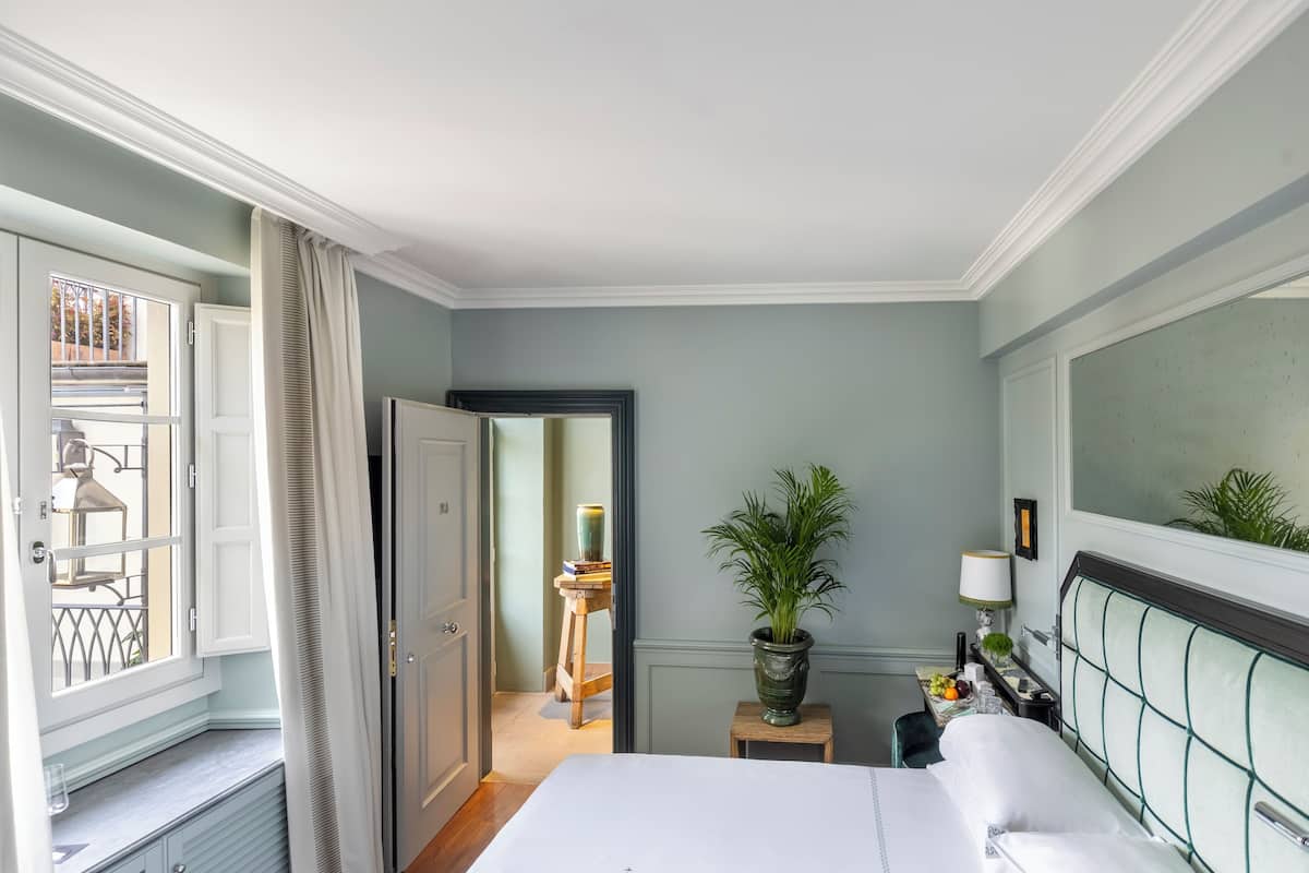 Habitación de hotel con cama, mesita de noche, planta en maceta, ventana con cortinas y vista al balcón exterior.