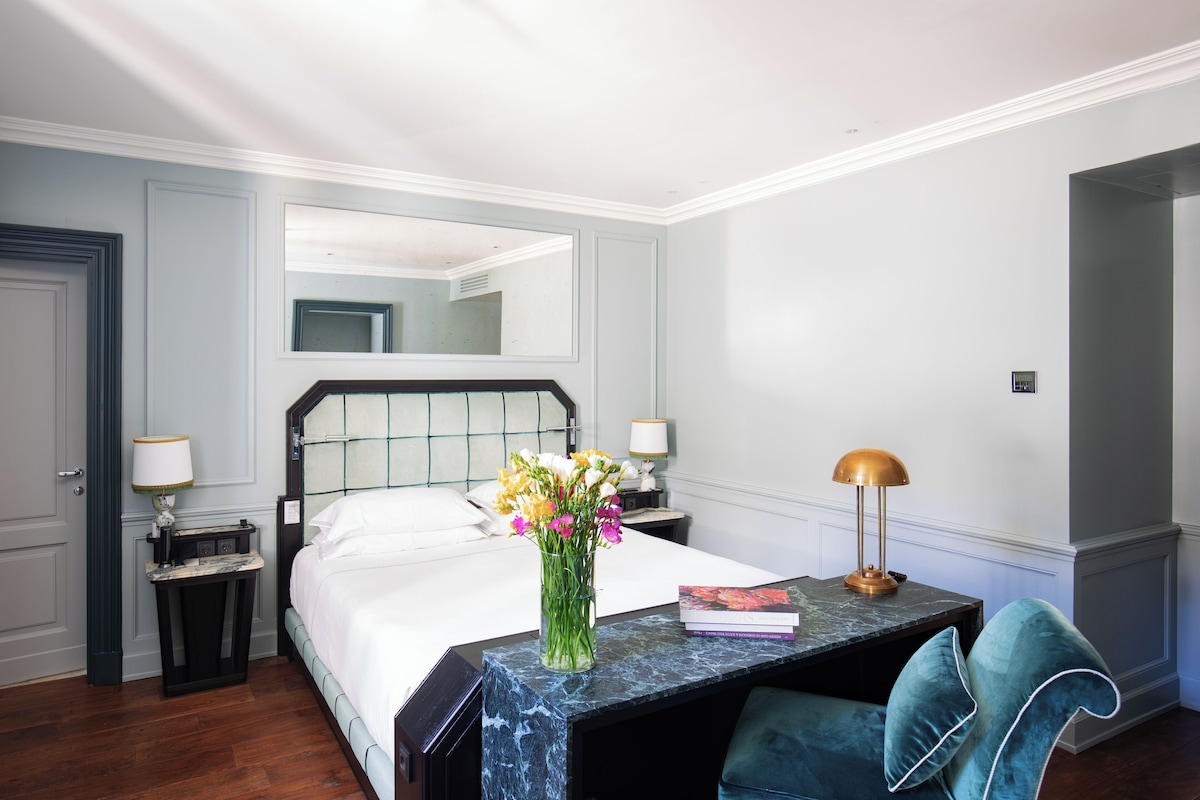 Habitación de hotel con cama, mesita de noche con lámpara, escritorio con lámpara, una silla y un jarrón con flores.