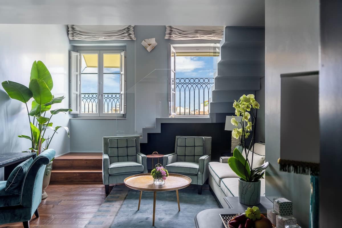 Una sala de estar moderna con piso de madera, una mesa redonda de centro y un sofá. Hay dos ventanas con balcones, y se ve una escalera.