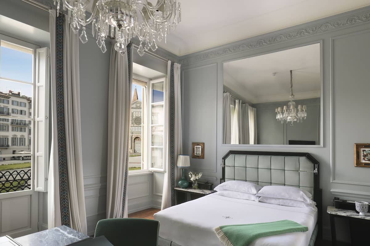 Habitación de hotel con una cama grande, un candelabro, vistas de un edificio a través de la ventana y un espejo en la pared.