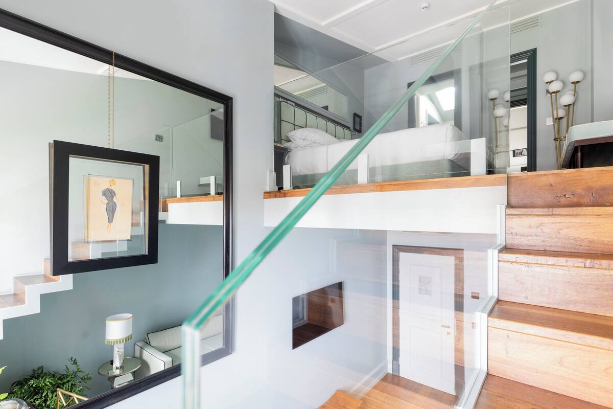 Una escalera moderna con barandillas de vidrio y escalones de madera que conduce a una zona en el segundo piso con una cama y una mesita de noche.