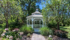 A gazebo with a white roof and open sides, surrounded by lush greenery and flowering plants. - Baymont by Wyndham Pompton Plains/Wayne (Pompton Plains)