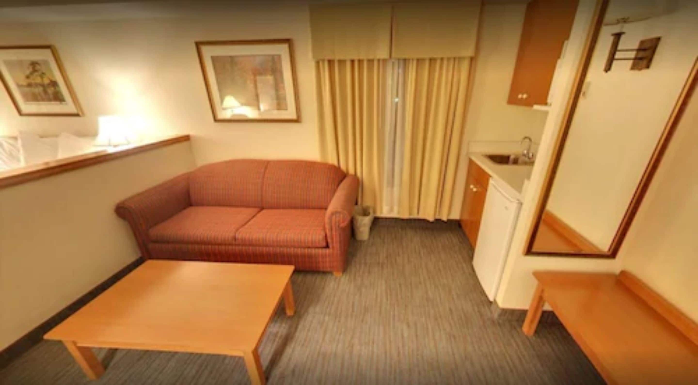 Quarto de hotel compacto com sofá, mesa pequena, cozinha compacta e janela com cortinas.